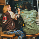 Two people sitting facing a bar cabinet, wearing embroidered SNL items. 