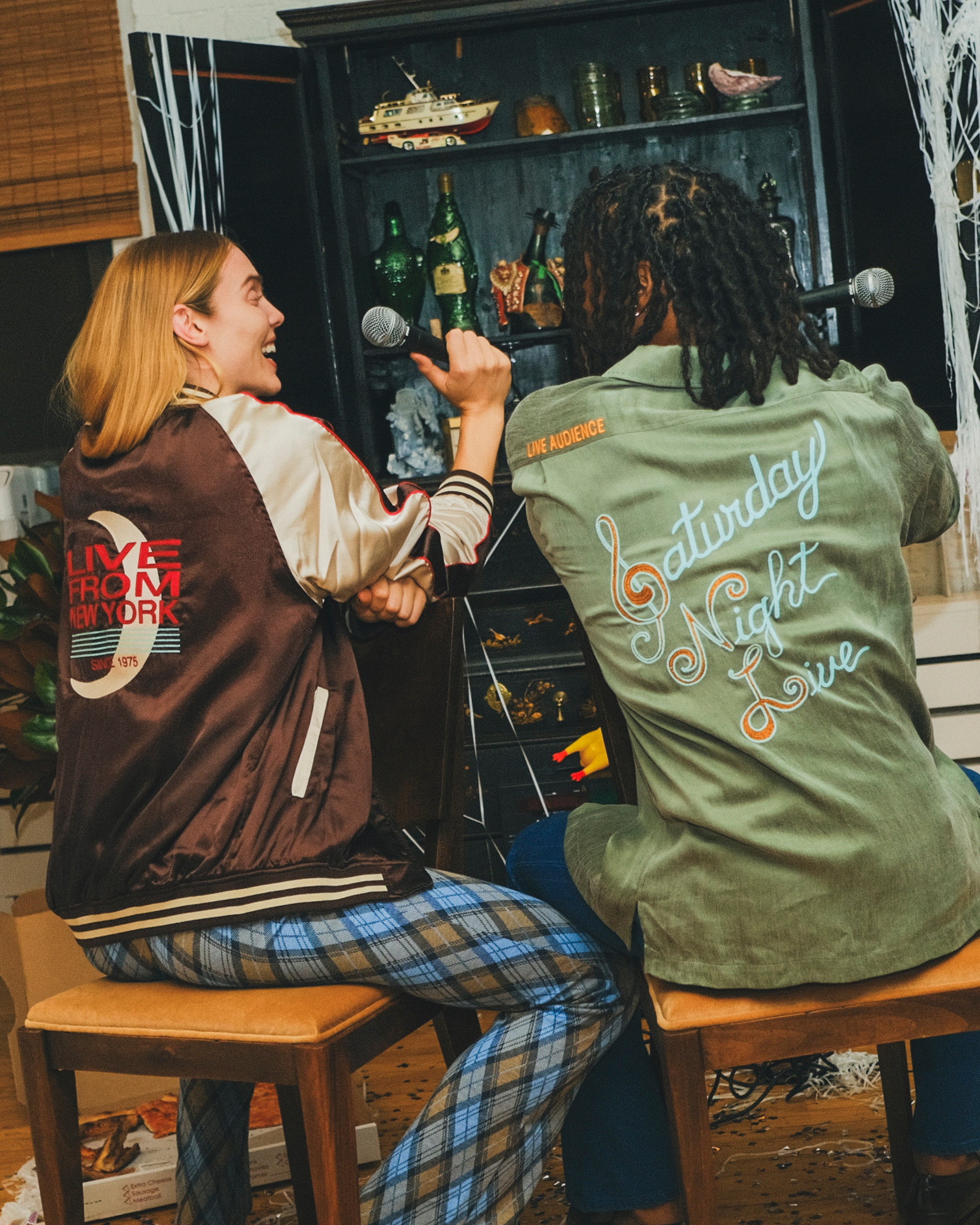 Two people sitting facing a bar cabinet, wearing embroidered SNL items. 