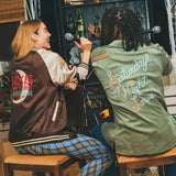 Two people sitting in front of a bar with decorative bottles, wearing embroidered SNL products. 