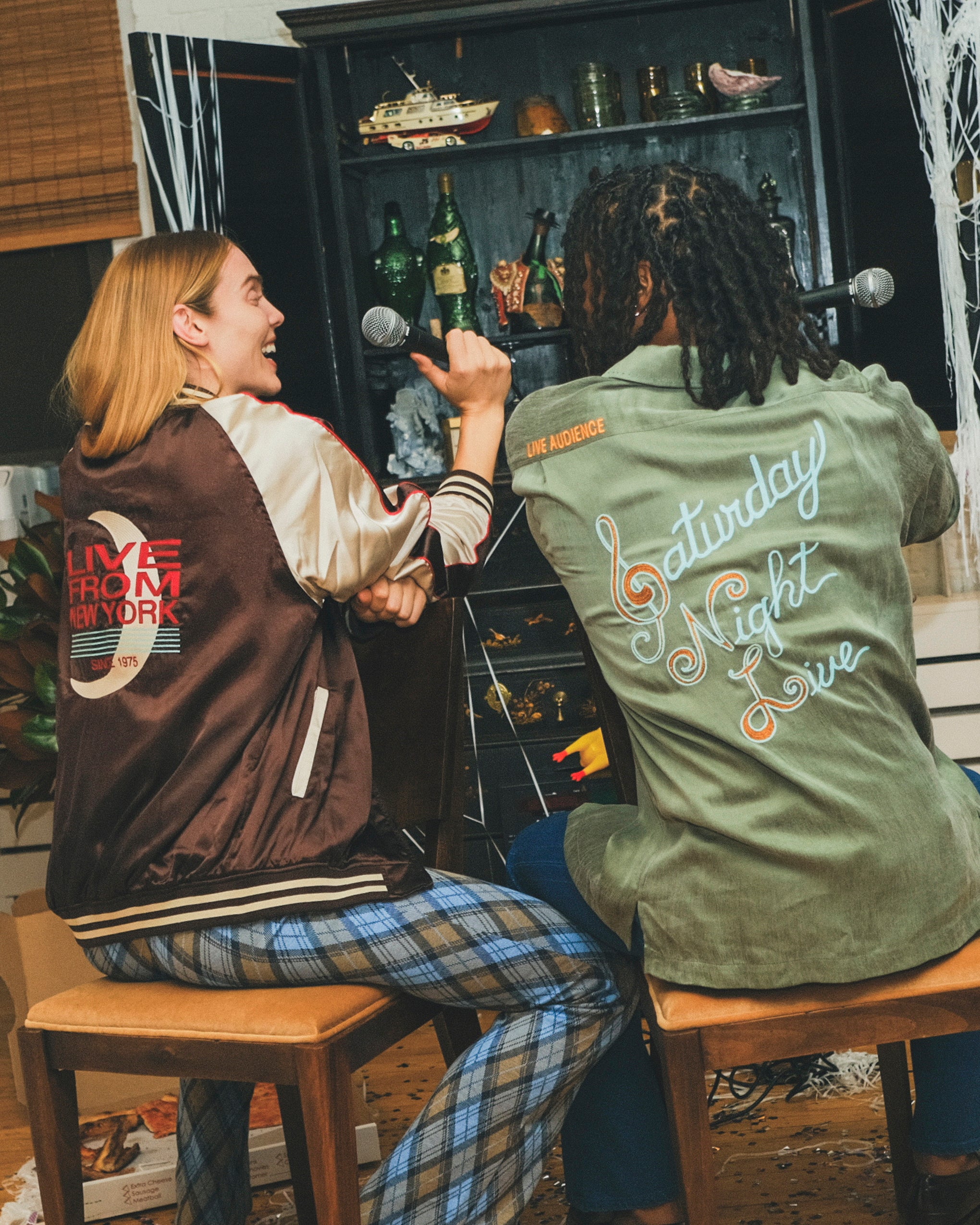 Two people sitting in front of a bar with decorative bottles, wearing embroidered SNL products. 