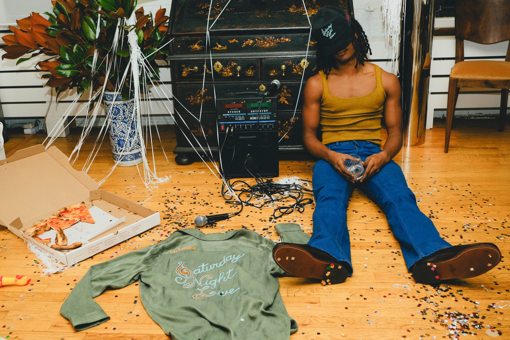 Person sitting on a wooden floor with scattered items including the green SNL shirt, pizza box, and a kaoroke machine.