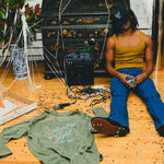 Person sitting on a wooden floor with scattered items including the green SNL shirt, pizza box, and a kaoroke machine.