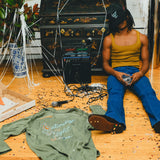 Person sitting on a wooden floor with scattered items including the green SNL shirt, pizza box, and a kaoroke machine.