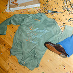Green long-sleeve shirt with 'Saturday Night Live' text on a wooden floor with confetti and a microphone.
