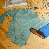 Green long-sleeve shirt with 'Saturday Night Live' text on a wooden floor with confetti and a microphone.