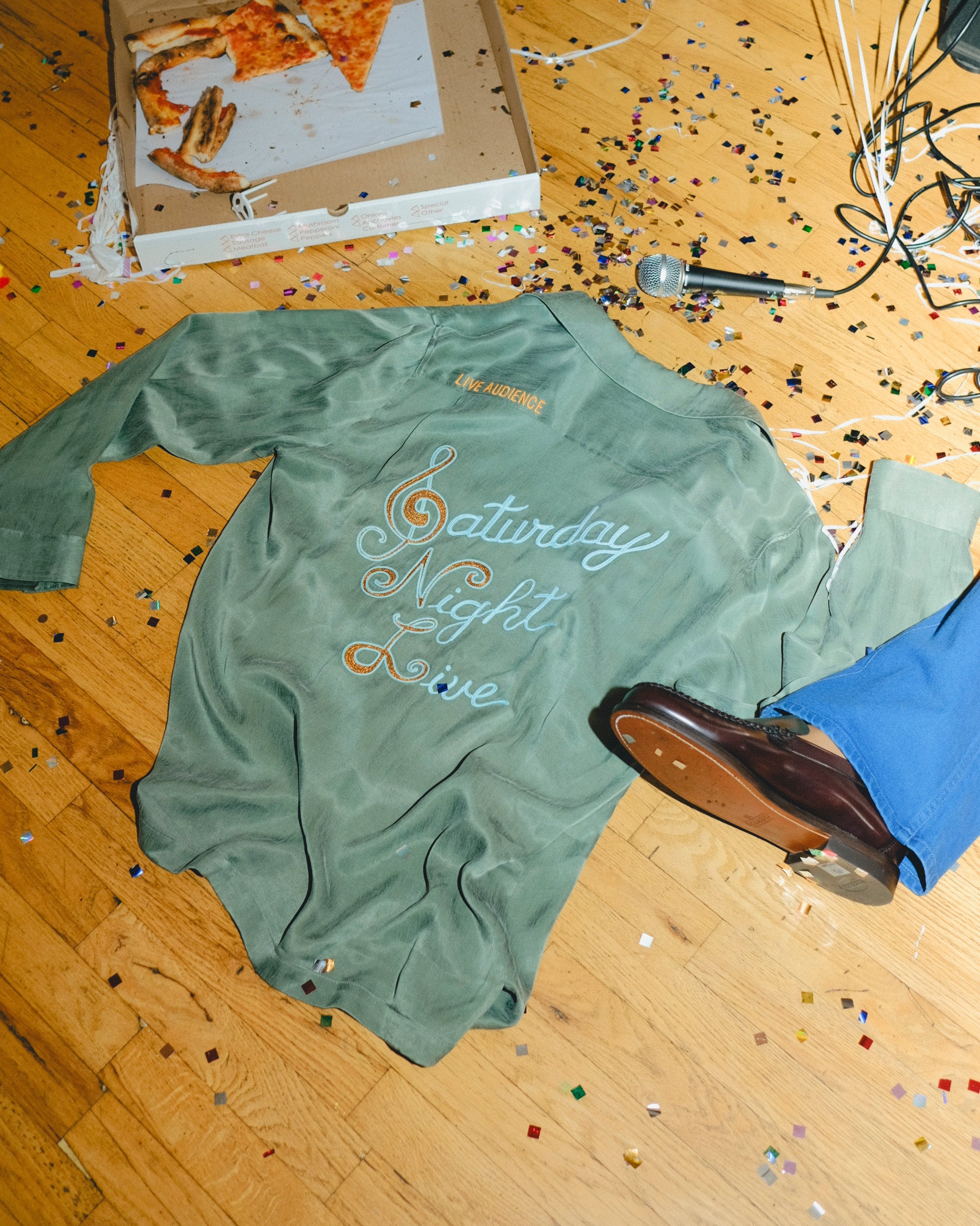 Green long-sleeve shirt with 'Saturday Night Live' text on a wooden floor with confetti and a microphone.