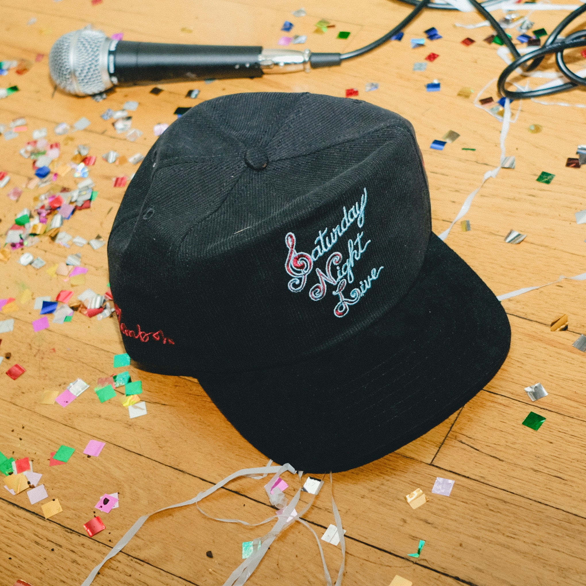 Black cap with 'Saturday Night Live' embroidery on a wooden floor with confetti and a microphone.