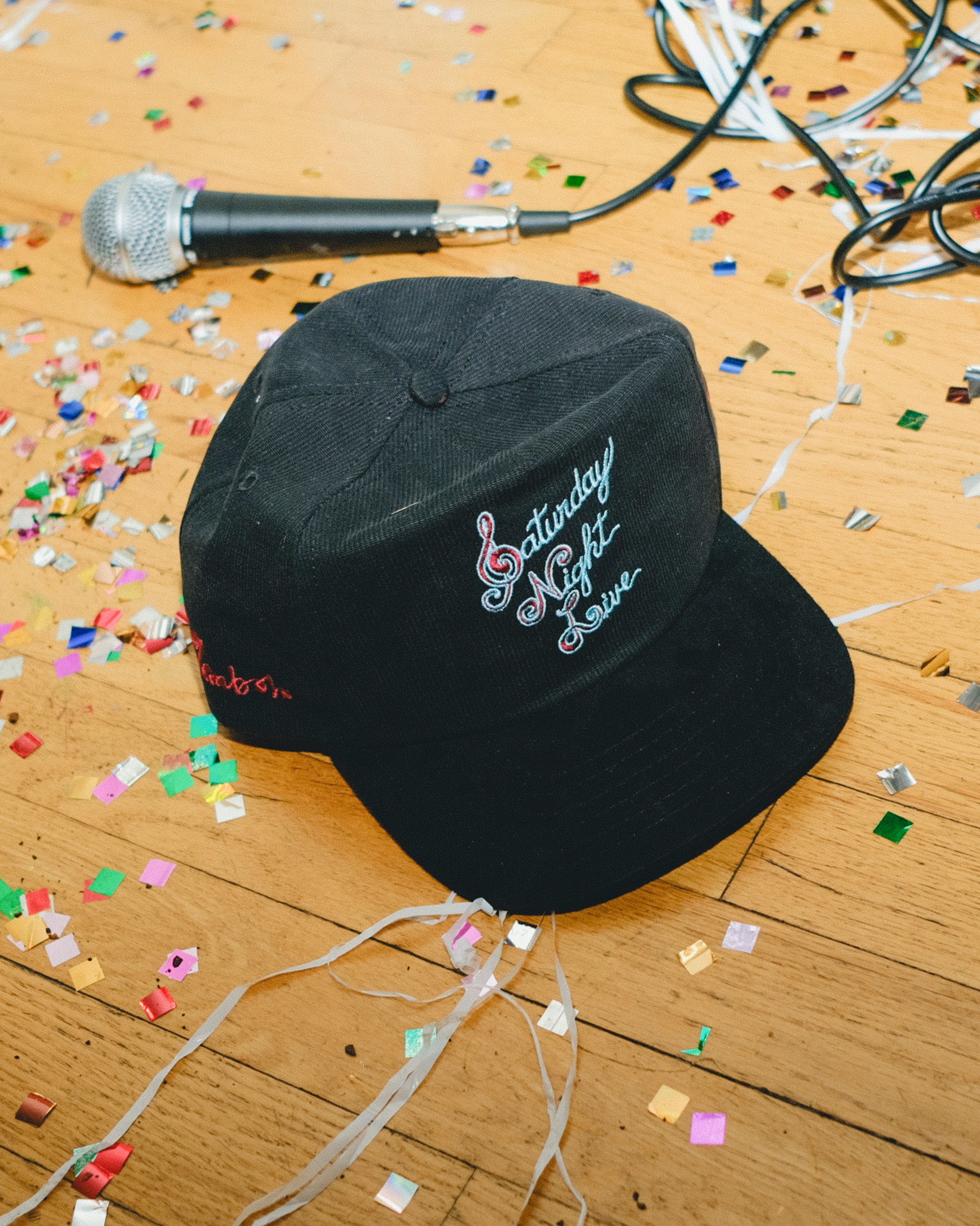 Black cap with 'Saturday Night Live' embroidery on a wooden floor with confetti and a microphone.
