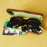 Lesca Lunetier X Tombolo lenses in Coral folded up and sitting inside of the brown suede case and tree-leaf patterned brown cleaning cloth that comes with the lenses, all positioned on a yellow background.