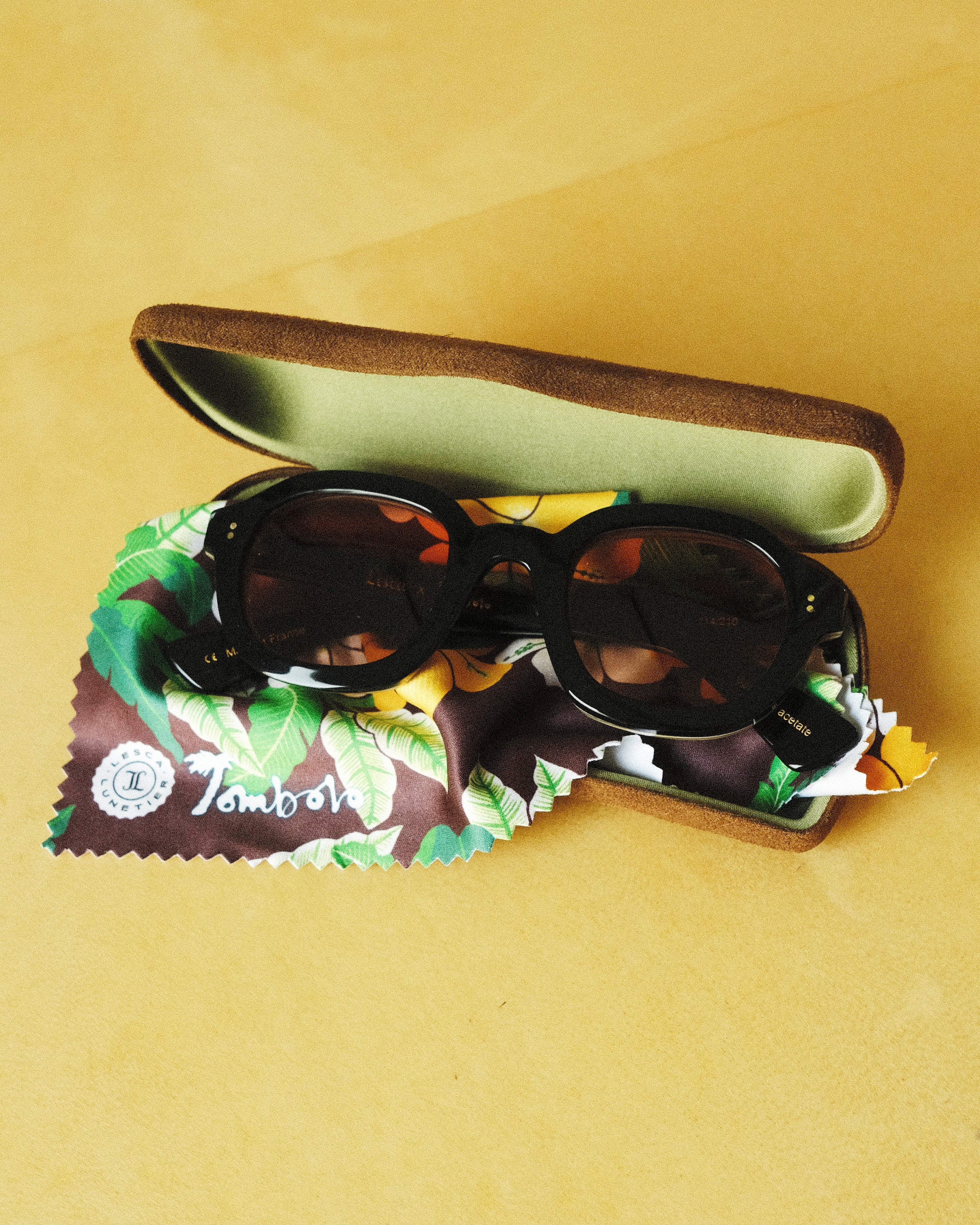 Lesca Lunetier X Tombolo lenses in Coral folded up and sitting inside of the brown suede case and tree-leaf patterned brown cleaning cloth that comes with the lenses, all positioned on a yellow background.