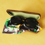 Lesca Lunetier X Tombolo lenses in Charcoal folded up and sitting inside of the brown suede case and tree-leaf patterned brown cleaning cloth that comes with the lenses, all positioned on a yellow background.