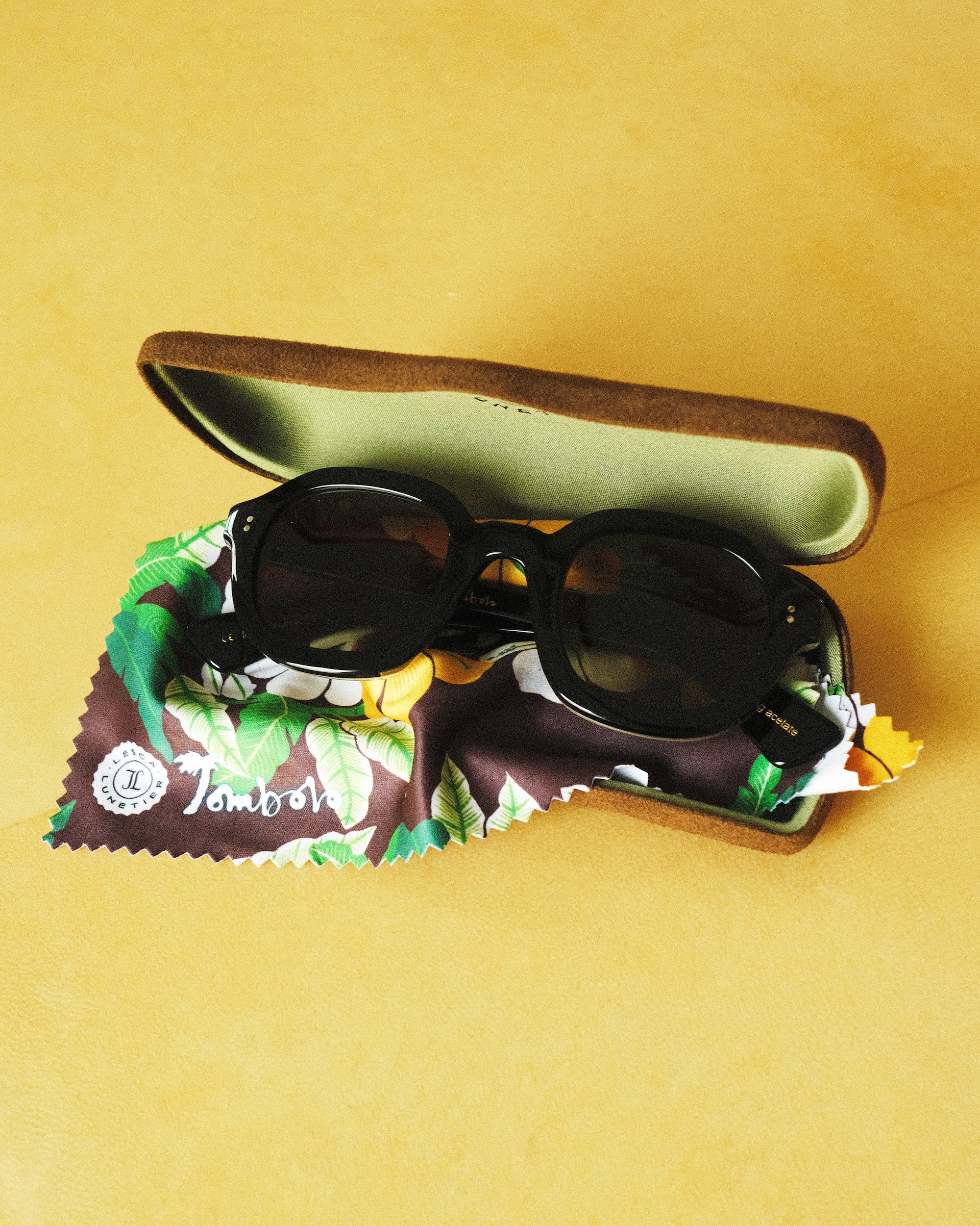 Lesca Lunetier X Tombolo lenses in Charcoal folded up and sitting inside of the brown suede case and tree-leaf patterned brown cleaning cloth that comes with the lenses, all positioned on a yellow background.