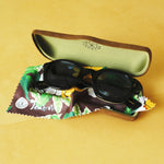 Lesca Lunetier X Tombolo Azure Lenses inside of the brown suede Lesca Lunetier X Tombolo glasses case sitting on top of the green and yellow tree/hibiscus patterned brown lense cloth.