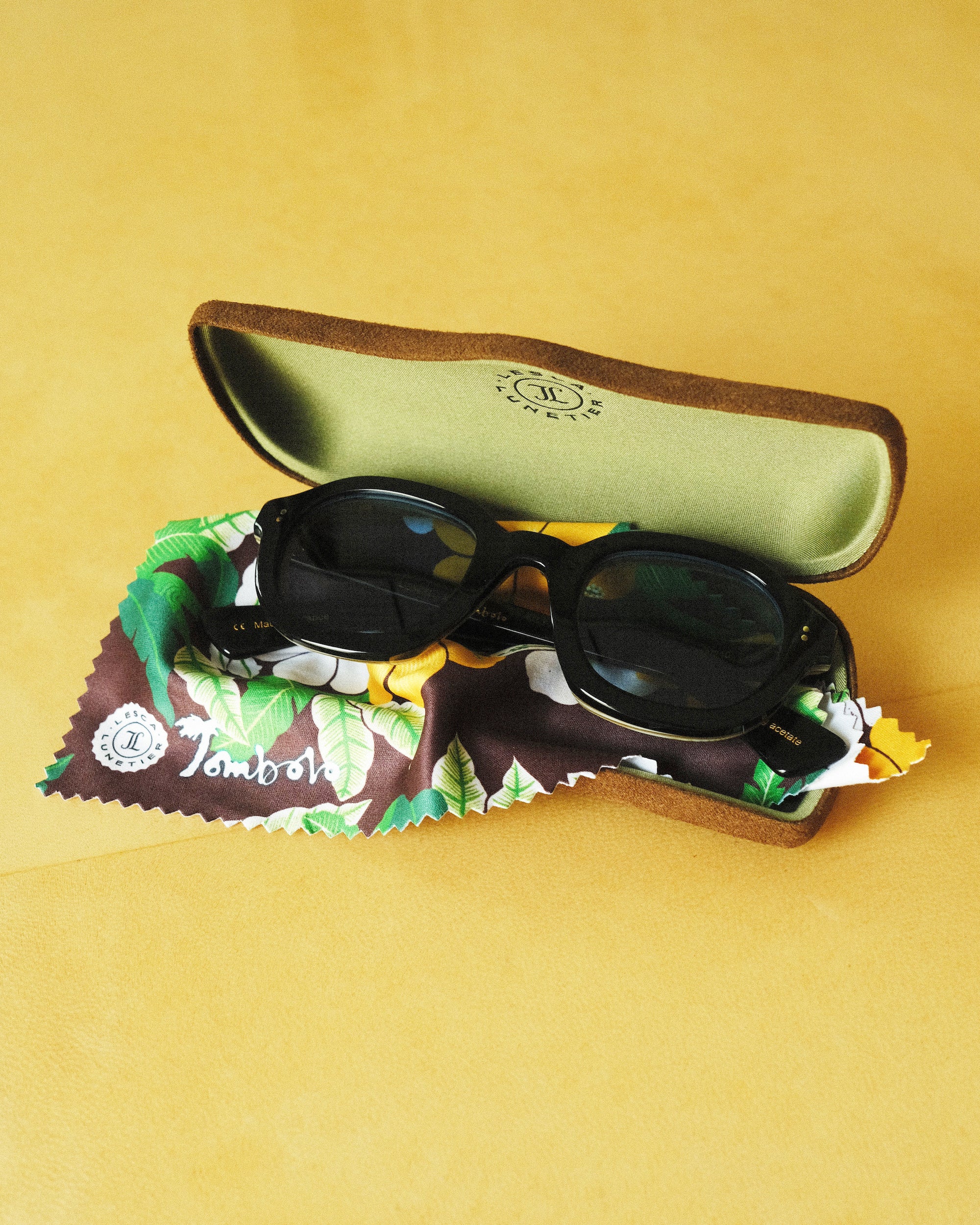 Lesca Lunetier X Tombolo Azure Lenses inside of the brown suede Lesca Lunetier X Tombolo glasses case sitting on top of the green and yellow tree/hibiscus patterned brown lense cloth.