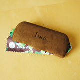 Lense case for Lesca Lunetier X Tombolo lenses that features a brown sueded case with script saying "lesca" in addition to a cleaning Fabric with both Lesca and Tombolo logos and a brown color with leaves printed on it throughout, all on a yellow background.