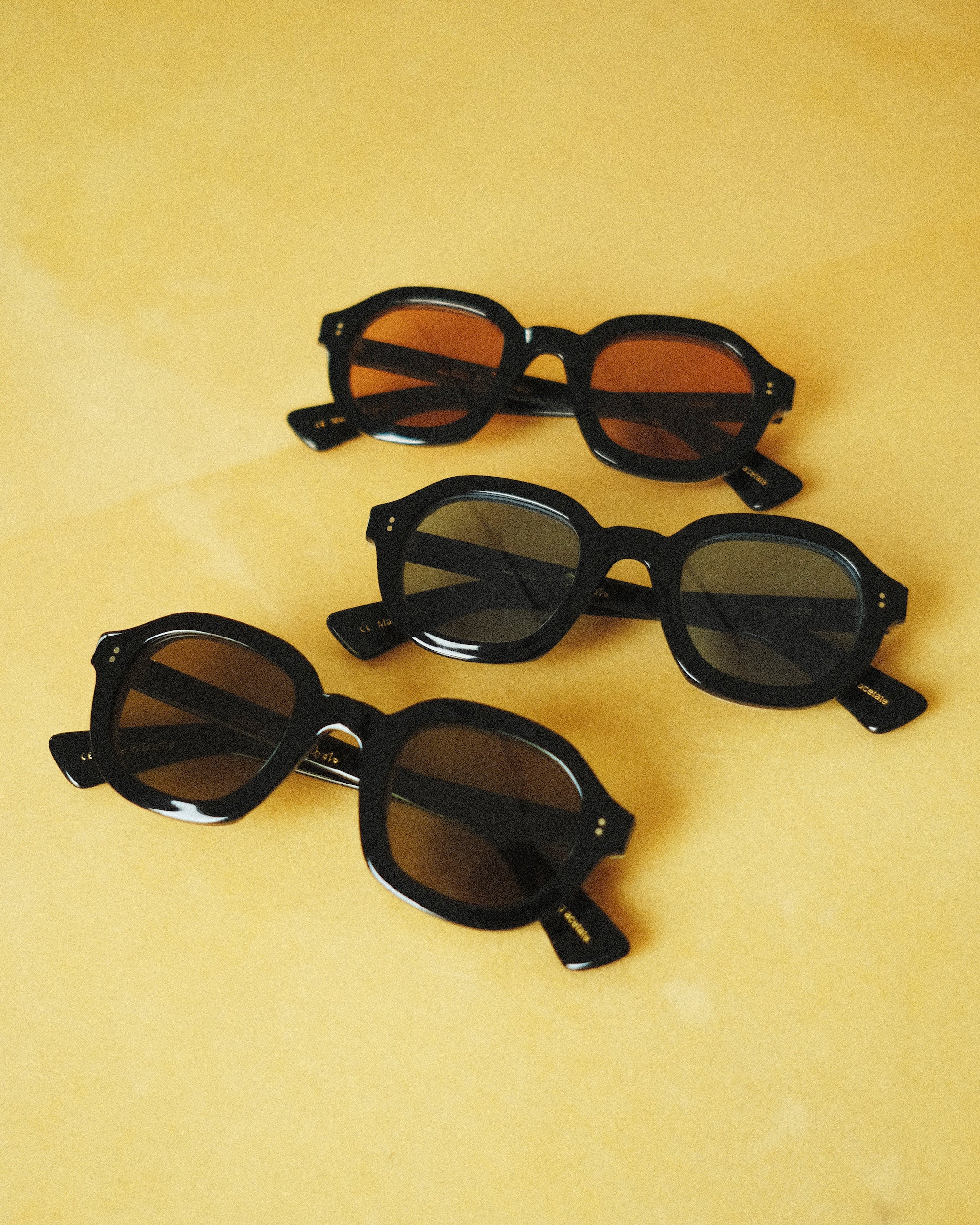three black-framed sunglasses against a yellow background; the first pair is black framed with pink lenses, the second pair is black framed with blue lenses, the third is black framed with black lenses