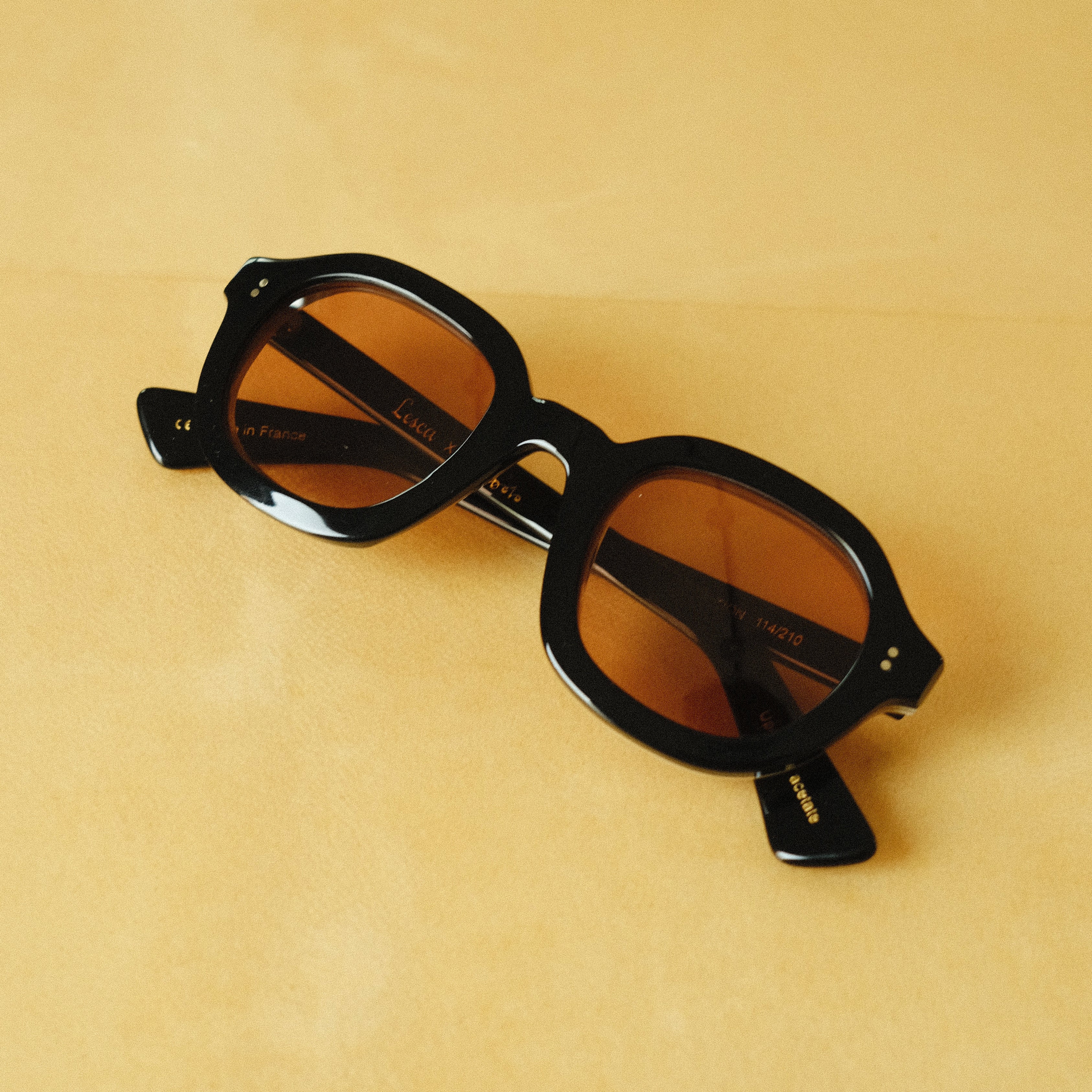 Lesca Lunetier X Tombolo front-facing product image with a black frame and squared round lenses in coral coloring on a yellow background.