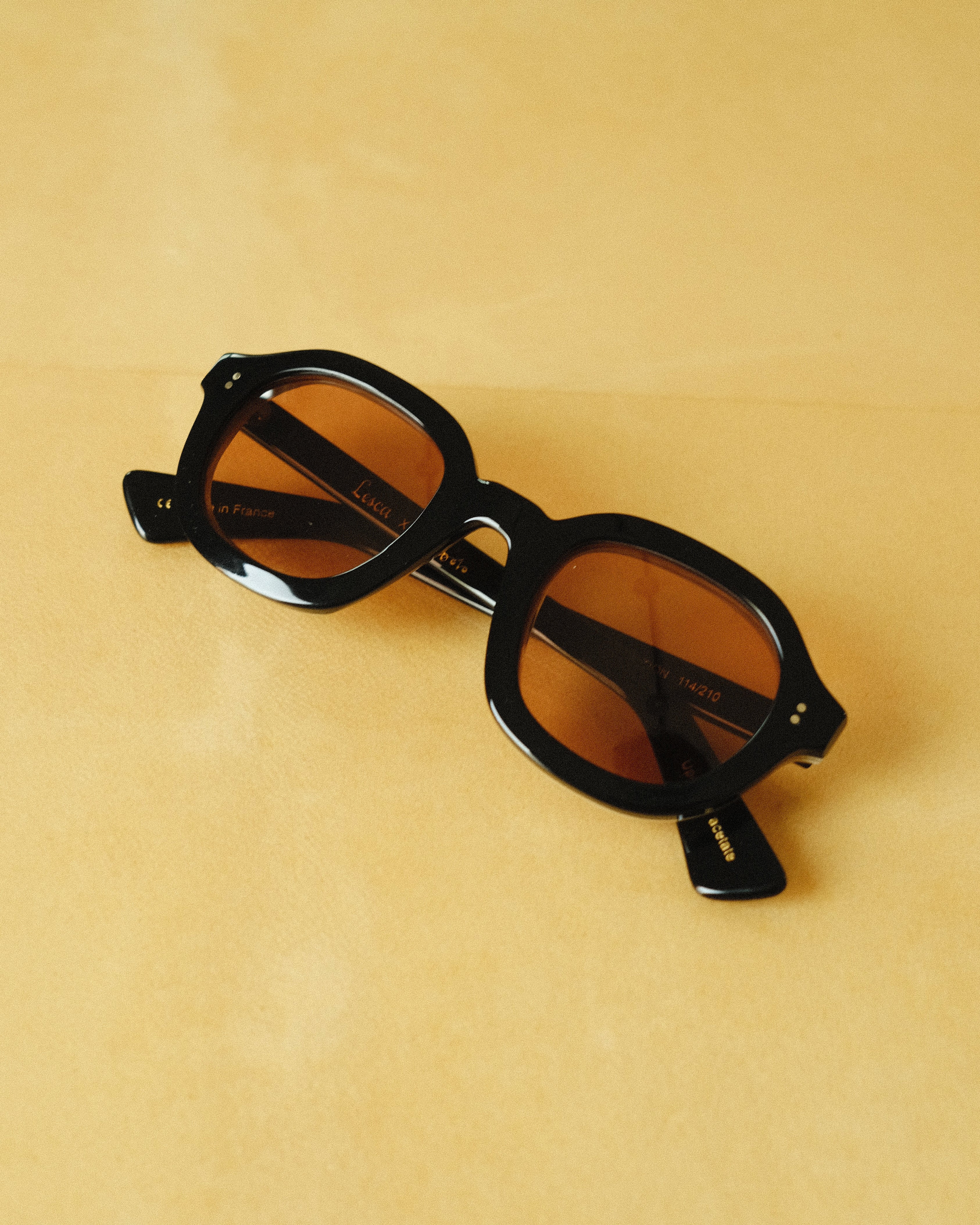 Lesca Lunetier X Tombolo front-facing product image with a black frame and squared round lenses in coral coloring on a yellow background.