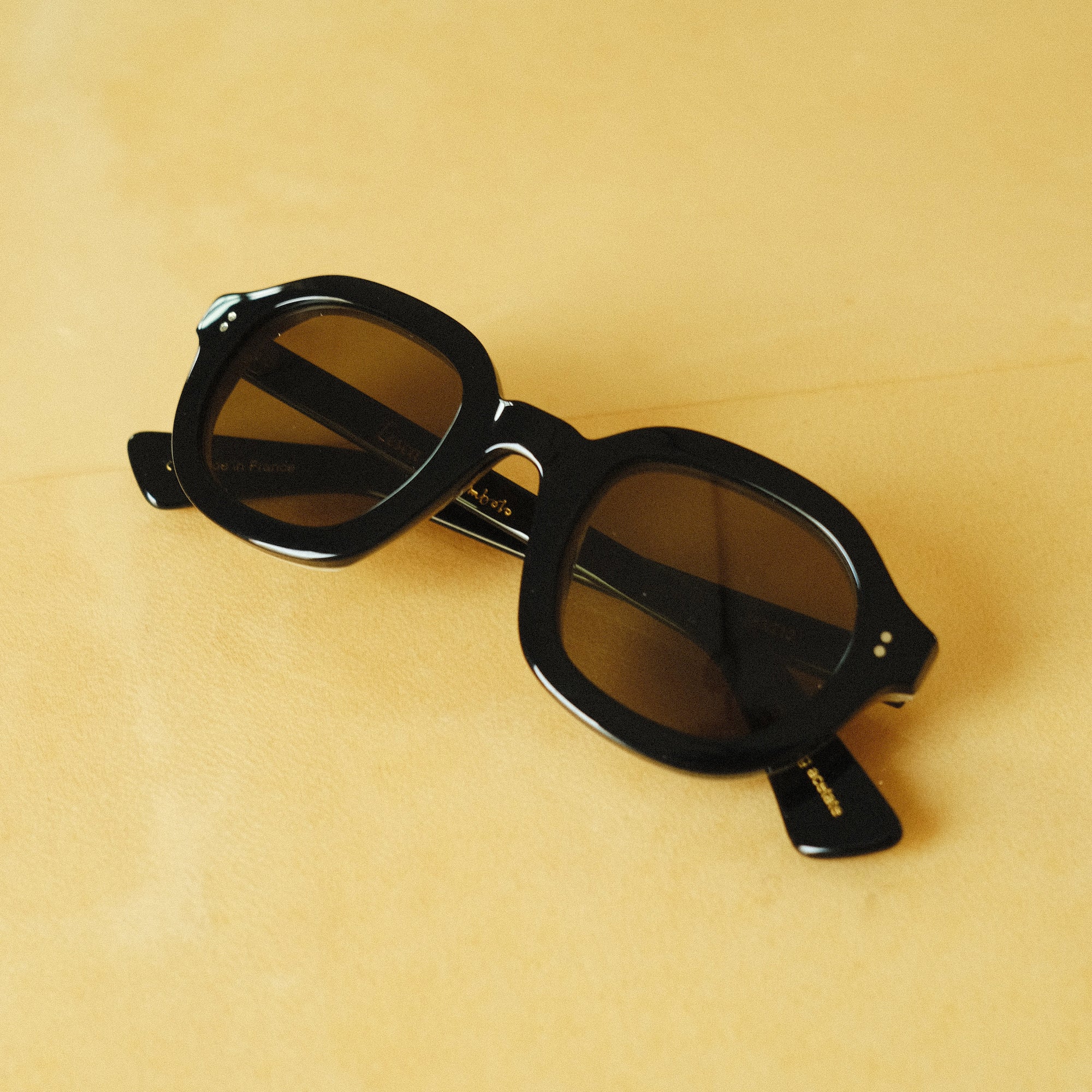 Front facing product image of Lesca Lunetier X Tombolo lenses in charcoal coloring with rounded square shaped lenses, folded up, on a yellow background