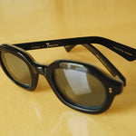 close up of thick, black framed sunglasses; Lesca x Tombolo interior etching visible; against a yellow background 