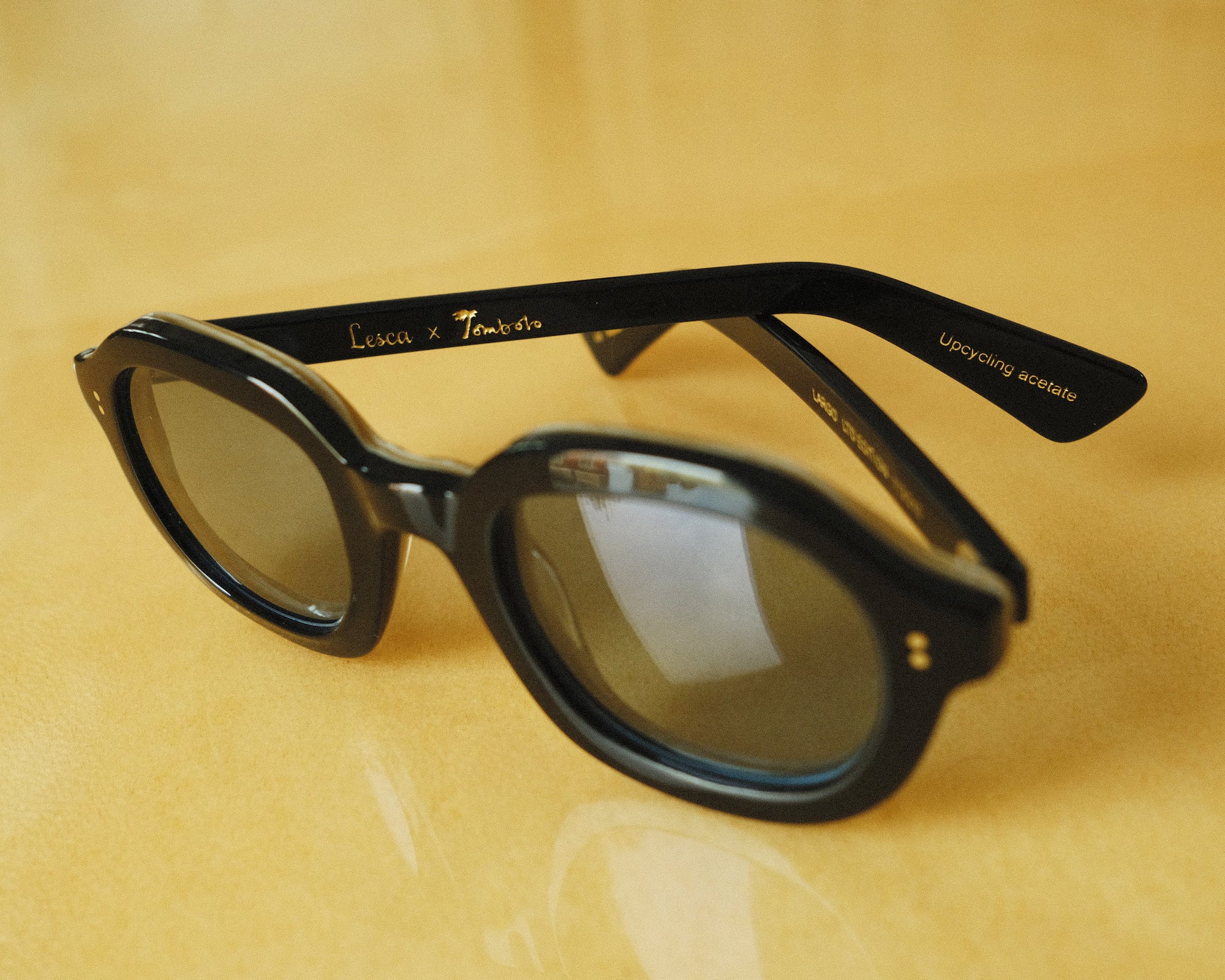 close up of thick, black framed sunglasses; Lesca x Tombolo interior etching visible; against a yellow background 