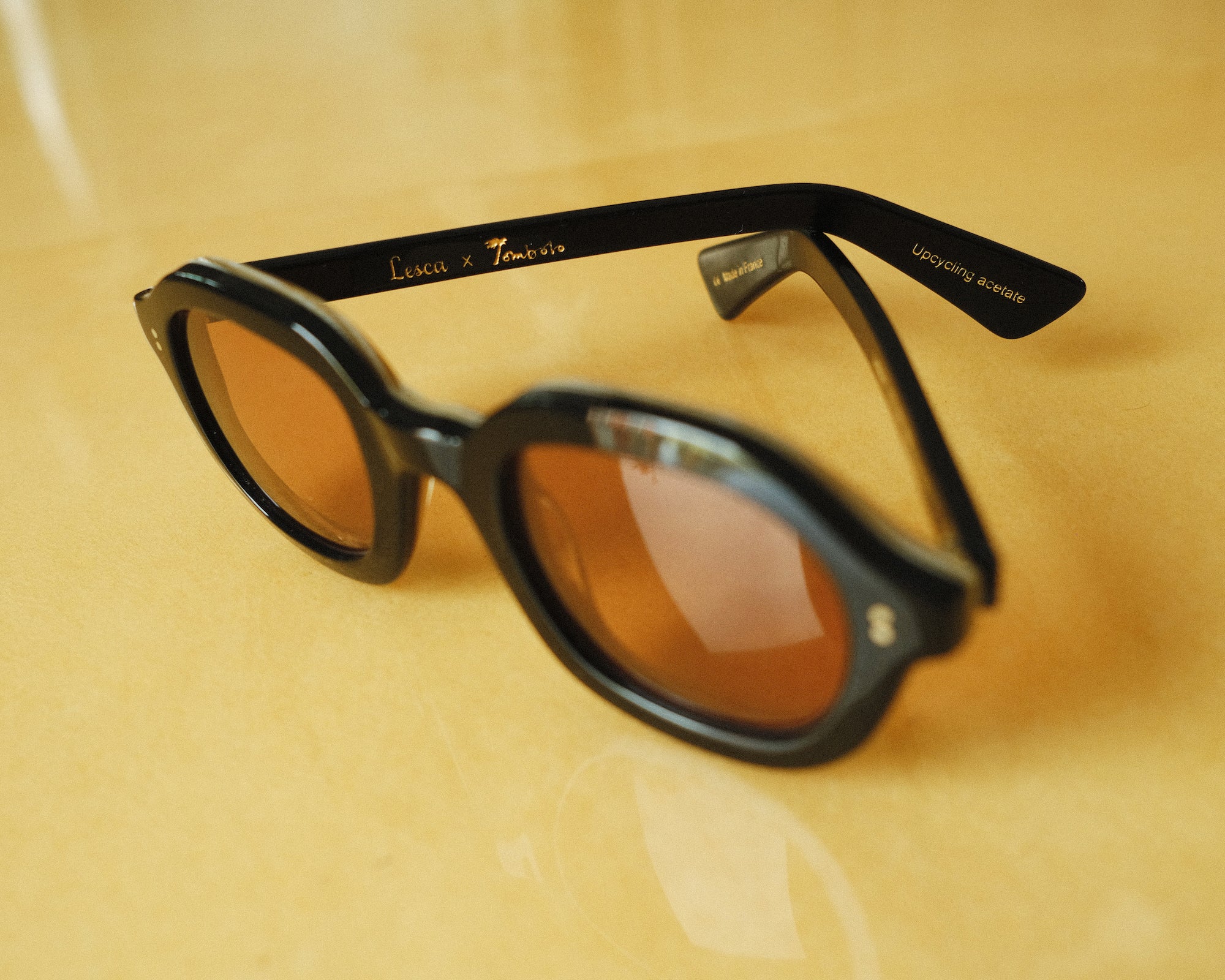 Lesca Lunetier X Tombolo lenses in Coral sideways-facing product image with emphasis on "Lesca x Tombolo" Logo graphics and 'upclycling acetate' denoting the types of material used to color the frame on a yellow background.
