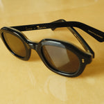 Lesca Lunetier X Tombolo lenses in charcoal sideways-facing product image with emphasis on "Lesca x Tombolo" Logo graphics and 'upclycling acetate' denoting the types of material used to color the frame on a yellow background.