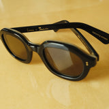 Lesca Lunetier X Tombolo lenses in charcoal sideways-facing product image with emphasis on "Lesca x Tombolo" Logo graphics and 'upclycling acetate' denoting the types of material used to color the frame on a yellow background.