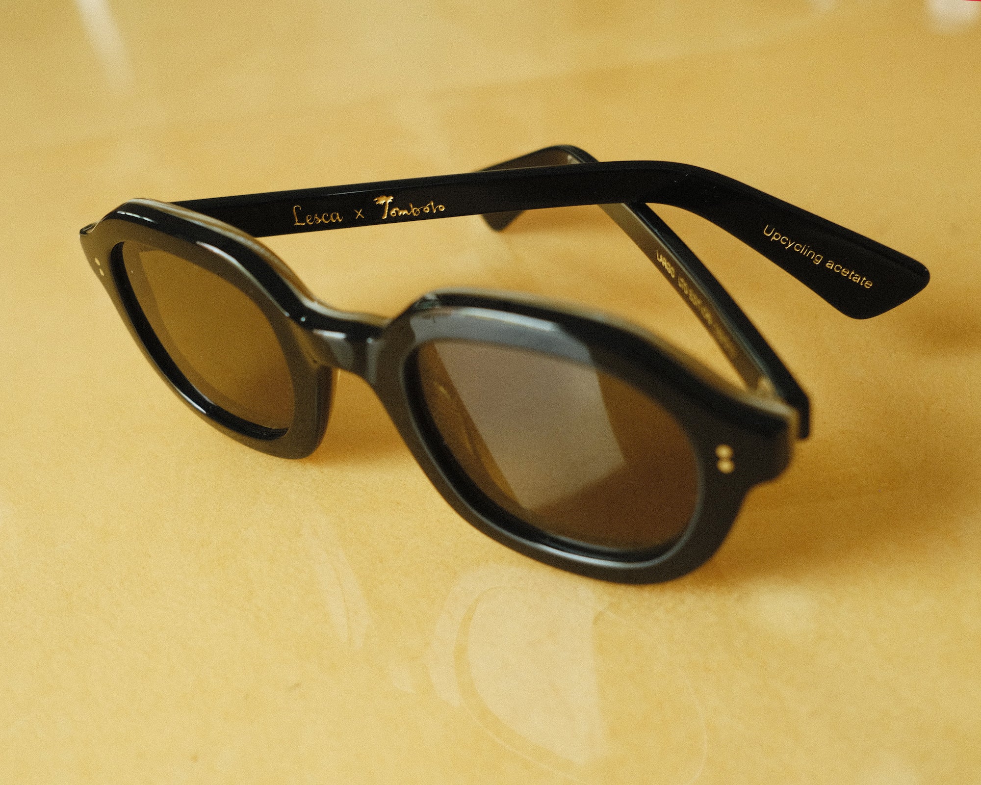 Lesca Lunetier X Tombolo lenses in charcoal sideways-facing product image with emphasis on "Lesca x Tombolo" Logo graphics and 'upclycling acetate' denoting the types of material used to color the frame on a yellow background.
