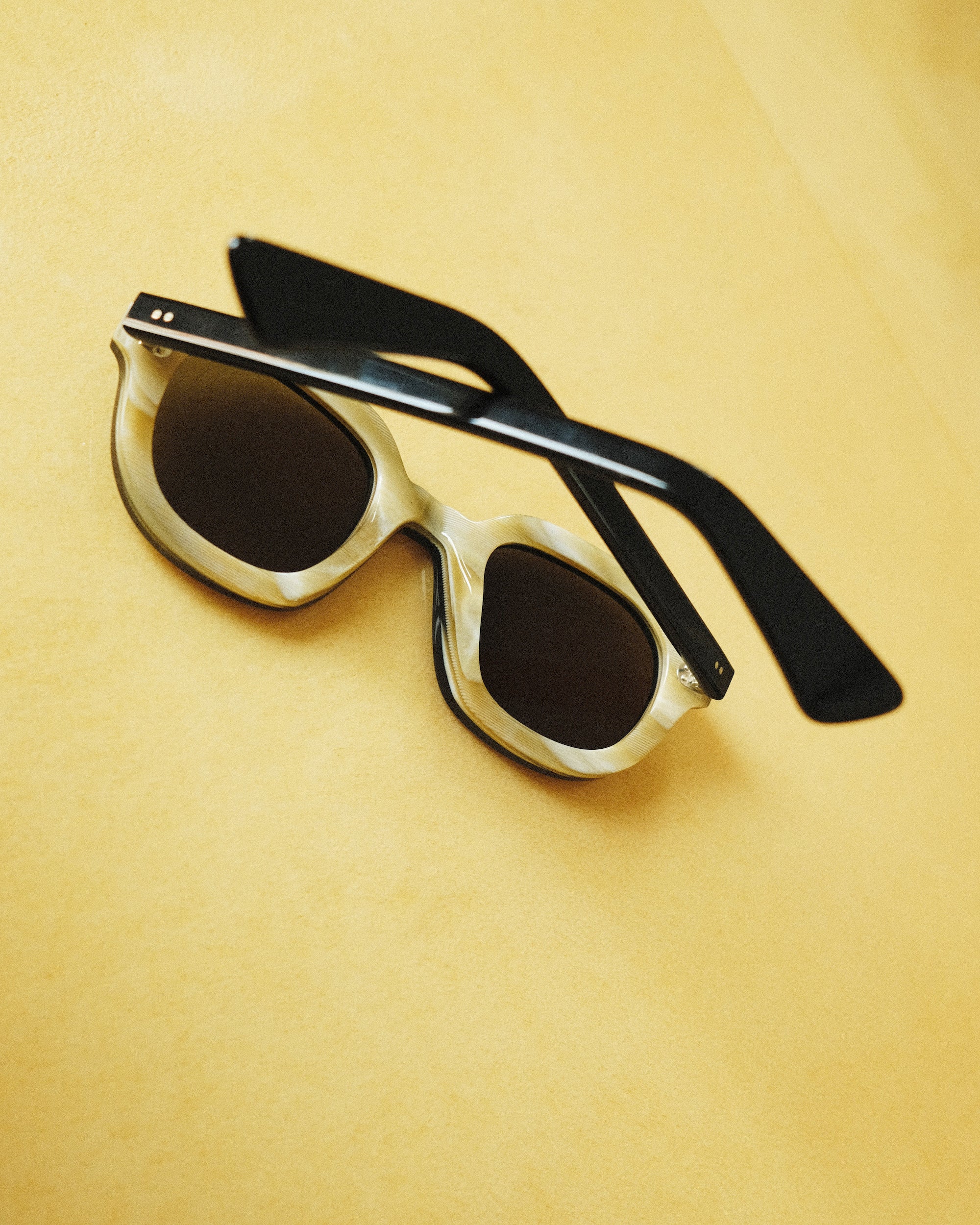 Lesca Lunetier X Tombolo lenses backwards-facing product image in charcoal coloring emphasizing the inner layer of horn-colored acetate with rounded square lenses on a yellow background.