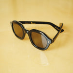 Lesca Lunetier X Tombolo lenses in charcoal using slightly angled product image where glasses lay half unfolded on a yellow background.