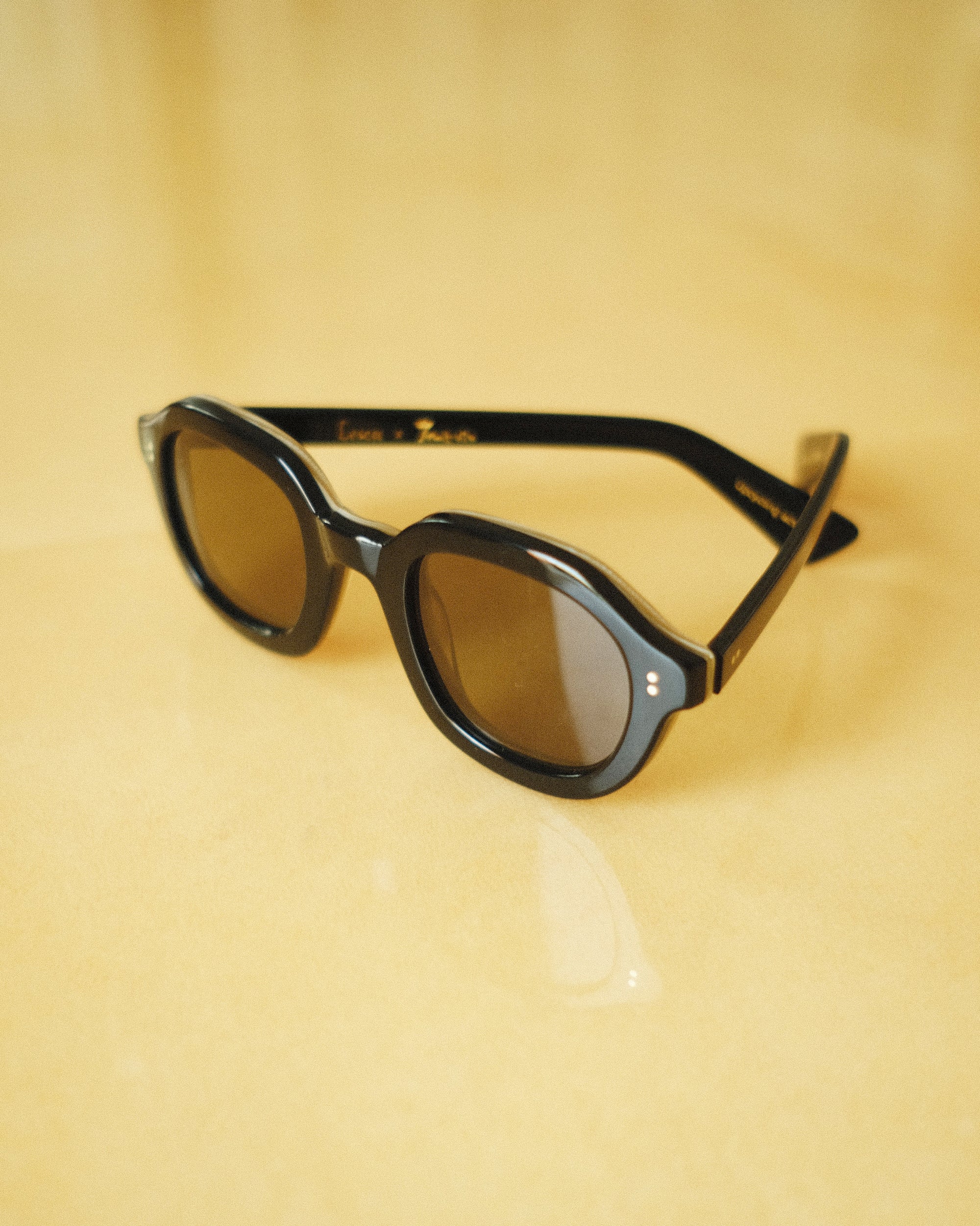 Lesca Lunetier X Tombolo lenses in charcoal using slightly angled product image where glasses lay half unfolded on a yellow background.