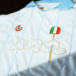 A close-up of a white and light blue pinstriped soccer jersey with gold embroidered 'una' lettering, a green-red-white circular crest, and an Italian flag patch.
