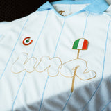 A close-up of a white and light blue pinstriped soccer jersey with gold embroidered 'una' lettering, a green-red-white circular crest, and an Italian flag patch.