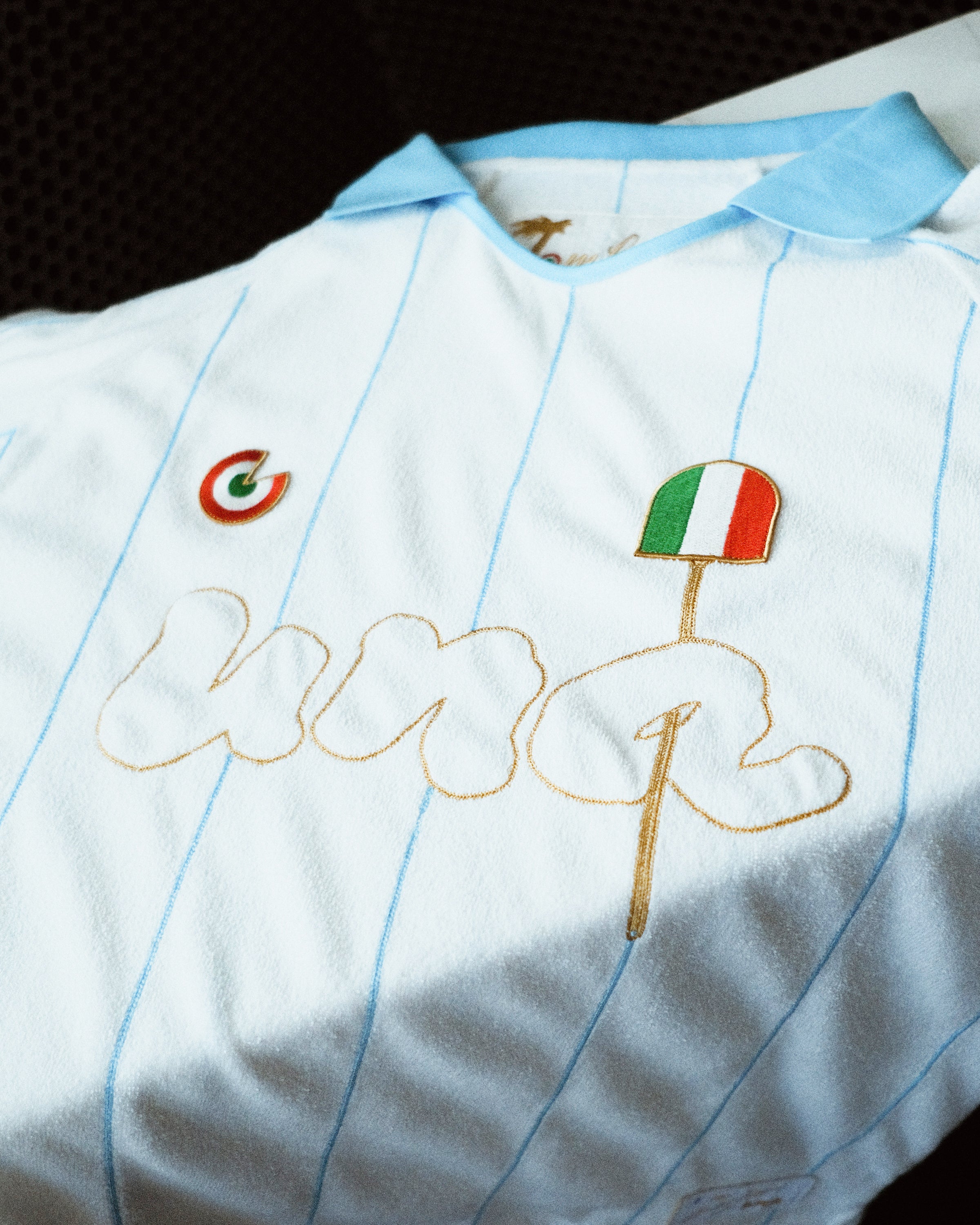 A close-up of a white and light blue pinstriped soccer jersey with gold embroidered 'una' lettering, a green-red-white circular crest, and an Italian flag patch.