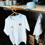 A white short-sleeve soccer jersey with light blue trim and embroidered details hangs on a wooden hanger in a shop or kitchen setting. A matching light blue cap and a white towel with blue and red stripes are also visible nearby.