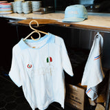 A white short-sleeve soccer jersey with light blue trim and embroidered details hangs on a wooden hanger in a shop or kitchen setting. A matching light blue cap and a white towel with blue and red stripes are also visible nearby.