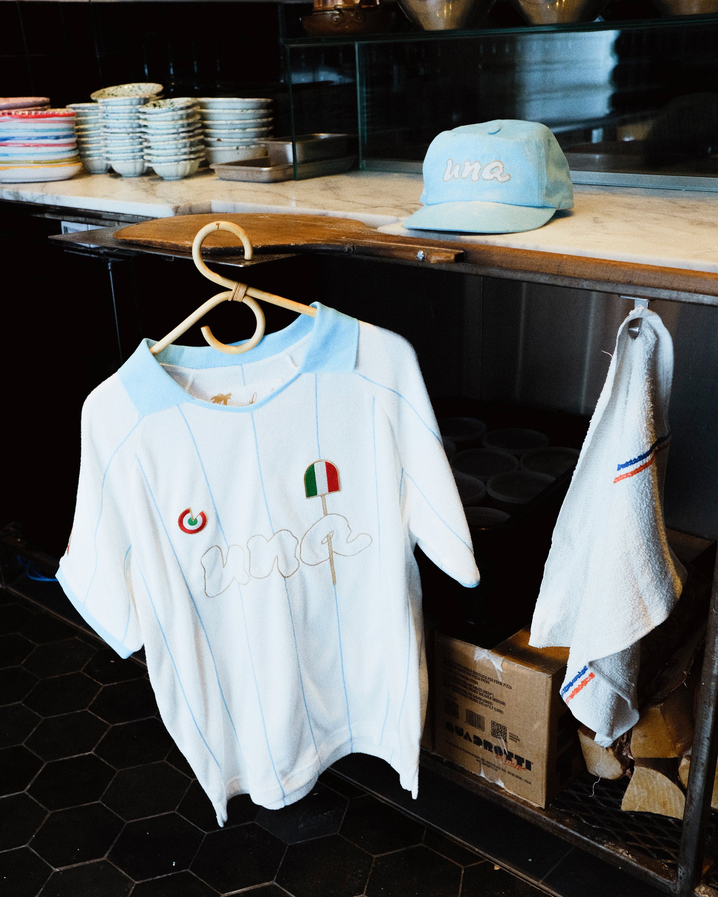 A white short-sleeve soccer jersey with light blue trim and embroidered details hangs on a wooden hanger in a shop or kitchen setting. A matching light blue cap and a white towel with blue and red stripes are also visible nearby.