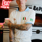 A tattooed man wearing a white pizza chef uniform with light blue trim, a matching light blue cap, and an apron stands in front of a tiled pizza oven. 