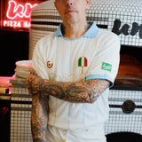 A tattooed man wearing a white pizza chef uniform with light blue trim, a matching light blue cap, and an apron stands in front of a tiled pizza oven. 
