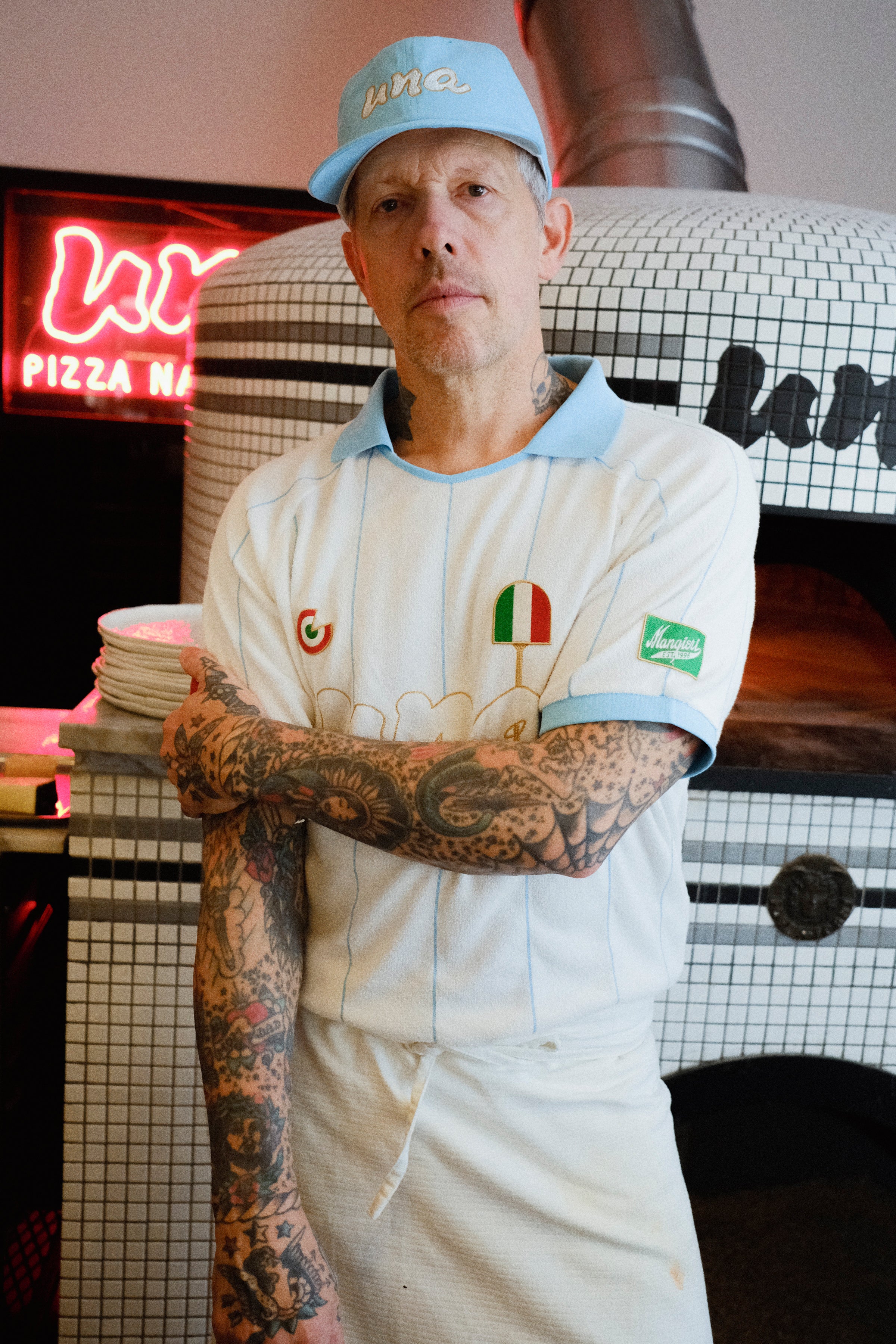 A tattooed man wearing a white pizza chef uniform with light blue trim, a matching light blue cap, and an apron stands in front of a tiled pizza oven. 