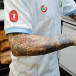A close-up of a person wearing a white soccer jersey with light blue pinstripes and sleeve trim. The jersey has multiple Italian patches.