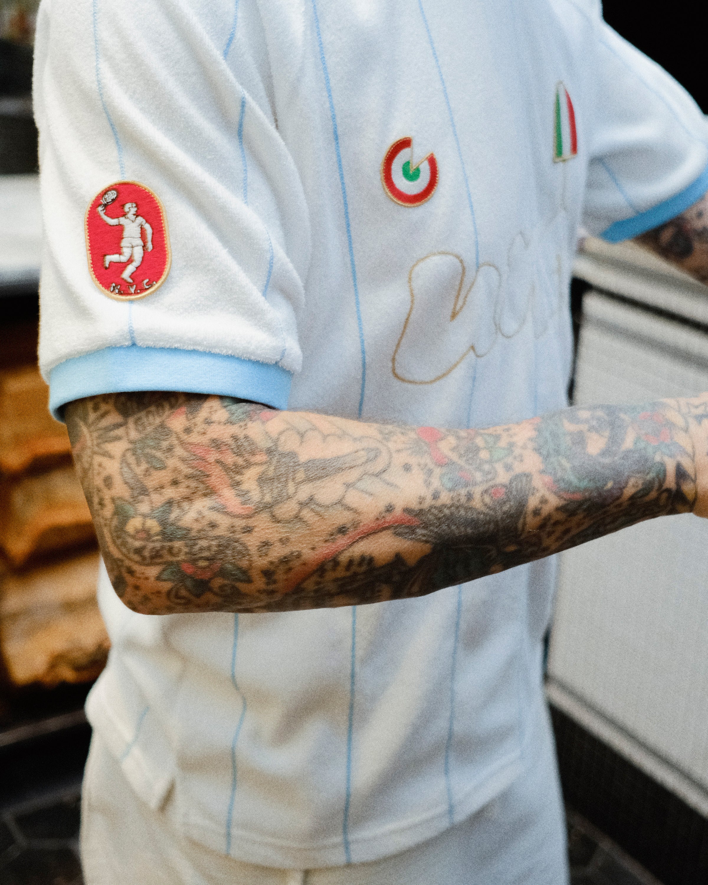 A close-up of a person wearing a white soccer jersey with light blue pinstripes and sleeve trim. The jersey has multiple Italian patches.