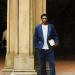 Man standing against a stone pillar wearing a navy blue corduroy jacket and pants with a white and navy striped shirt. He is holding two books and wearing brown loafers.