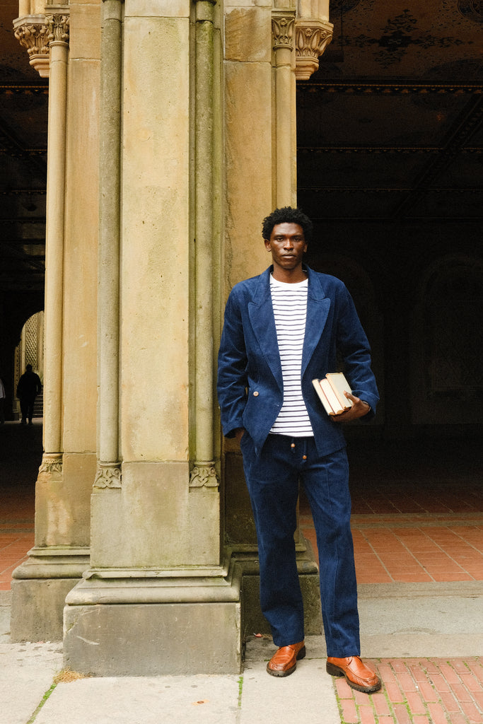 Man standing against a stone pillar wearing a navy blue corduroy jacket and pants with a white and navy striped shirt. He is holding two books and wearing brown loafers.