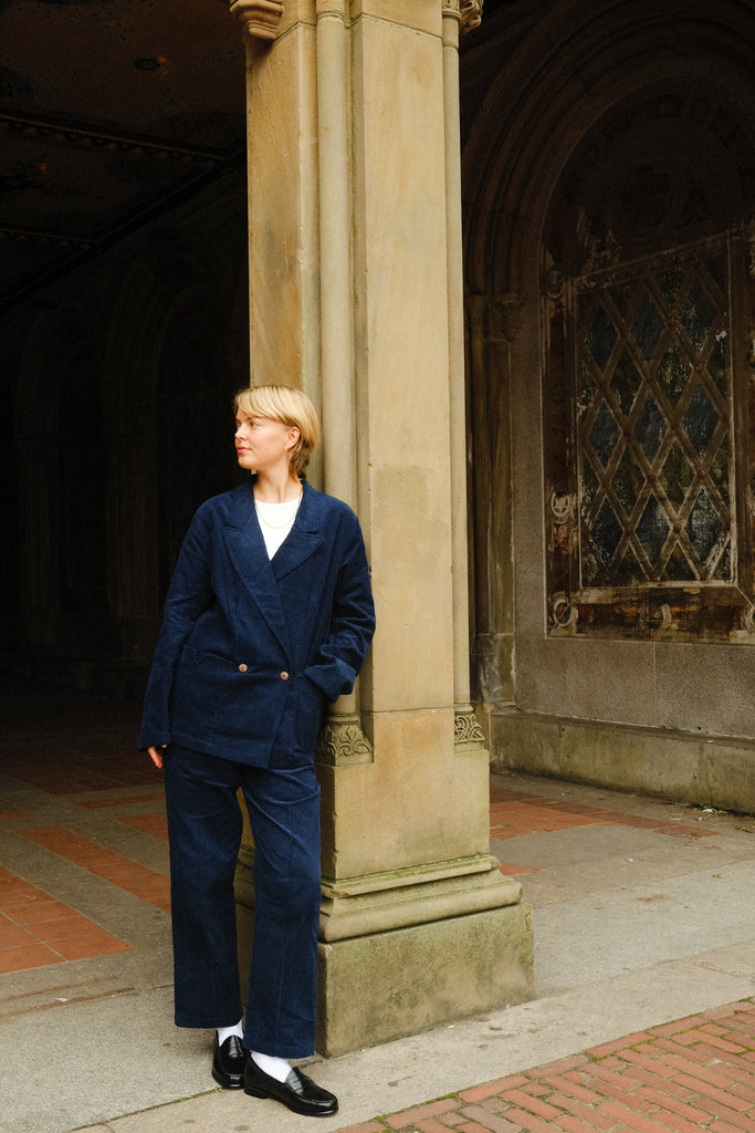 Woman, leaning against a pillar, wearing the full Navy Corduroy Easy Going Suit, with black loafers. 