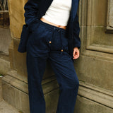 Close-up of a person wearing navy blue corduroy drawstring pants and a matching jacket, paired with a white cropped tank, white socks, black loafers, and a gold chain necklace.