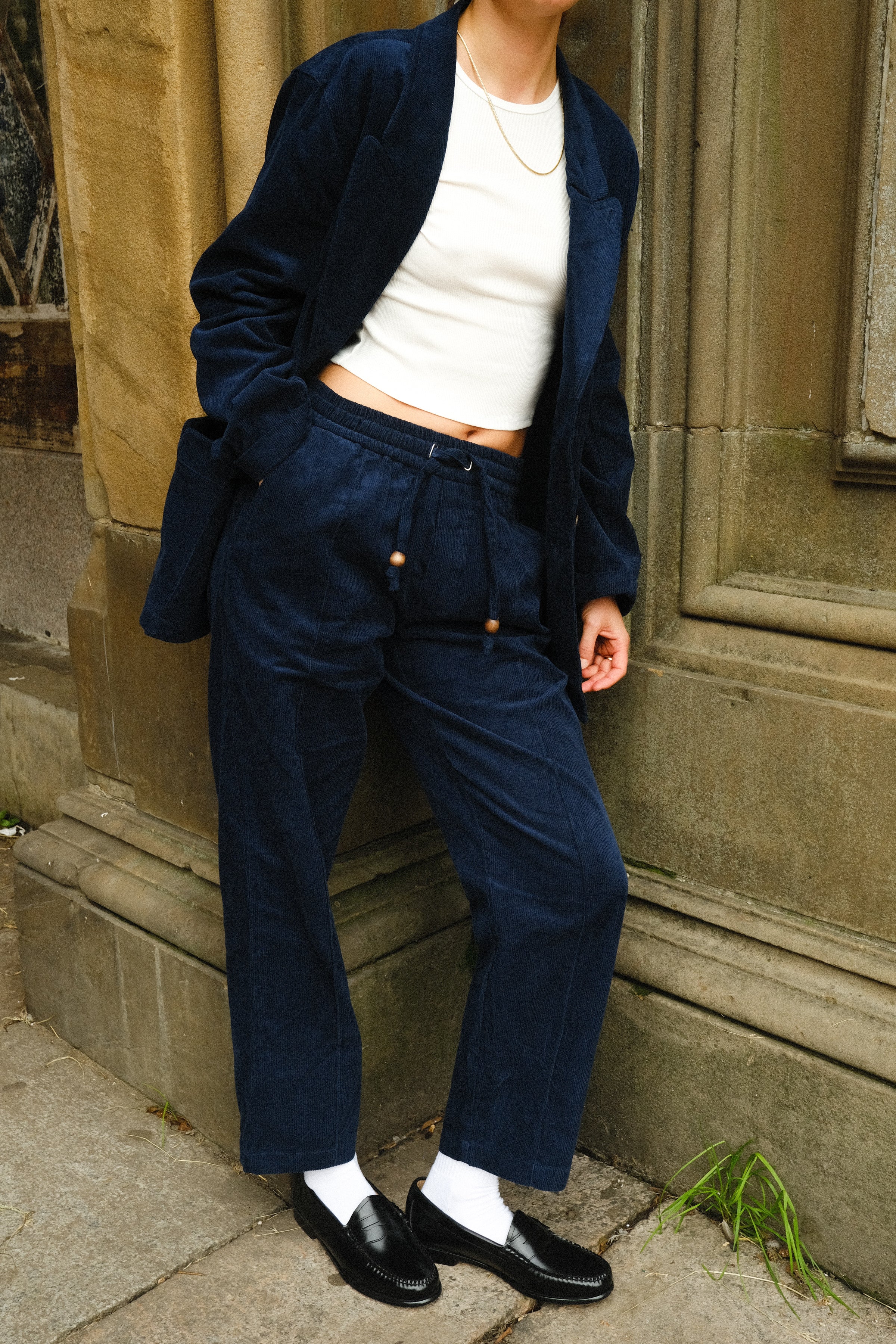 Close-up of a person wearing navy blue corduroy drawstring pants and a matching jacket, paired with a white cropped tank, white socks, black loafers, and a gold chain necklace.