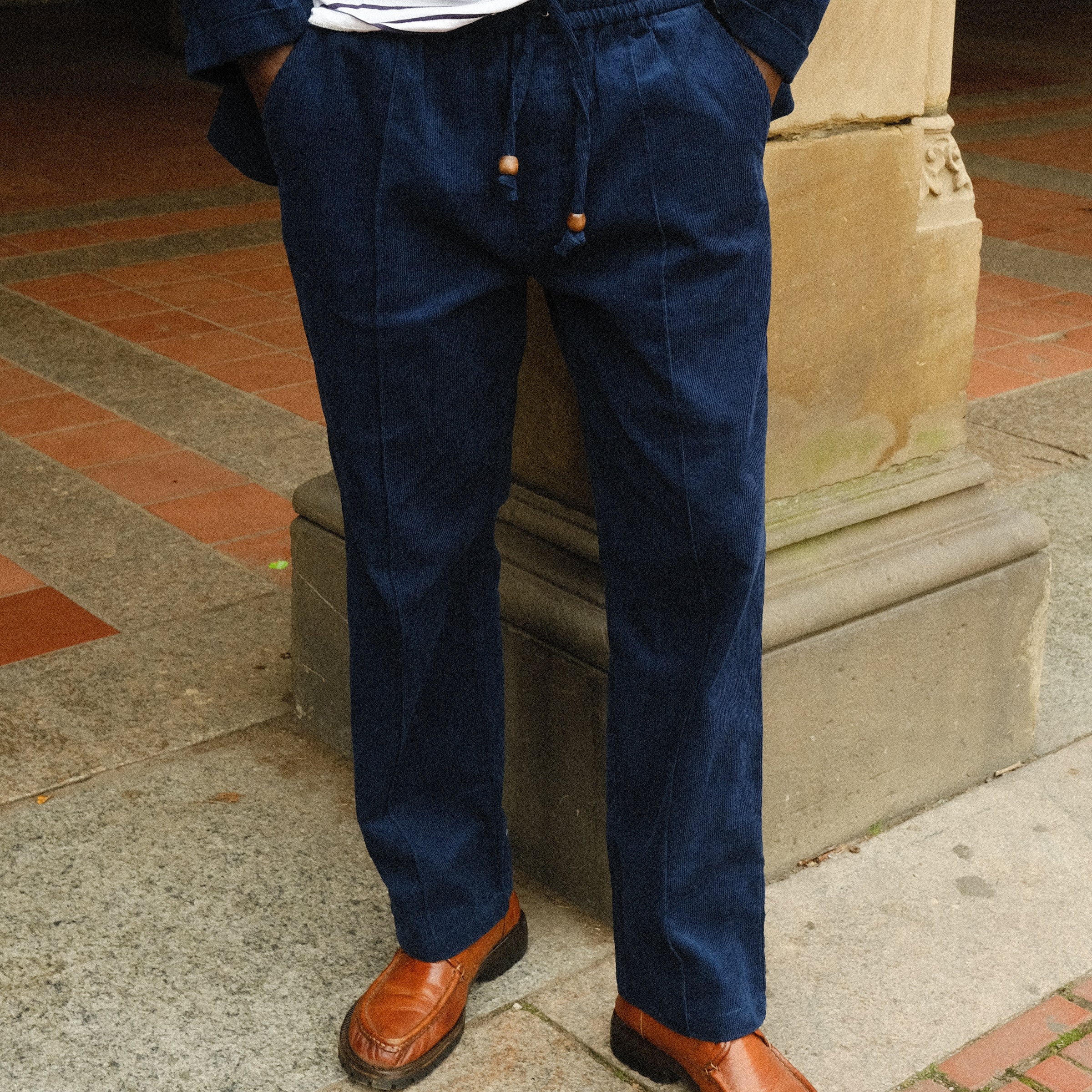 Close-up of a person wearing navy blue corduroy drawstring pants with a matching jacket, paired with a white and navy striped shirt and brown loafers.