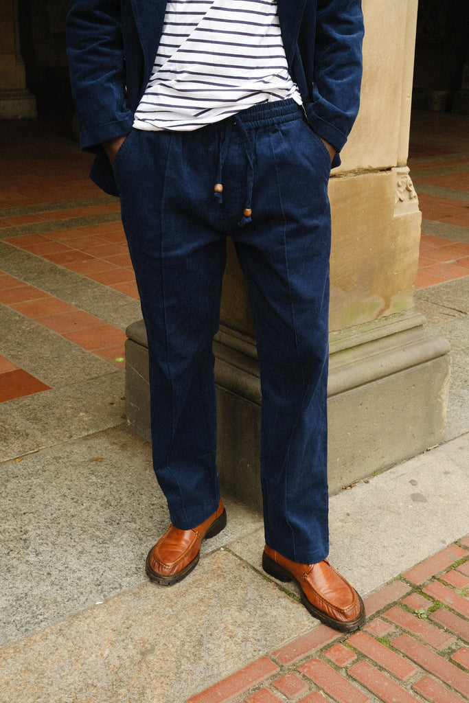 Close-up of a person wearing navy blue corduroy drawstring pants with a matching jacket, paired with a white and navy striped shirt and brown loafers.